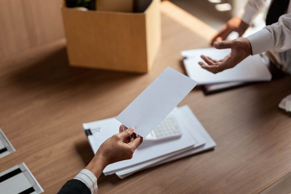 A desk featuring a hand handing off a paper, and a coworker accepting it. The desk also has papers, and a cardboard box, and other office items.